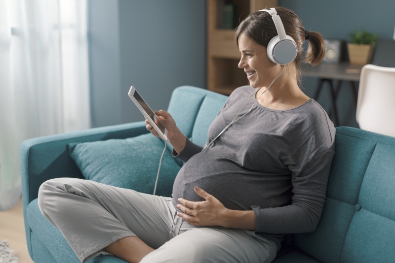 White woman sat holding stomach on a blue sofa, listening to music through her silver headphones