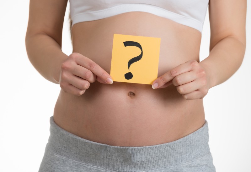 White woman holding small paper square with question mark over uncovered stomach.